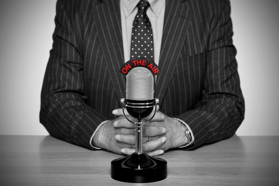 Photo of a news broadcaster sat at a desk and retro microphone with an On The Air illuminated sign.