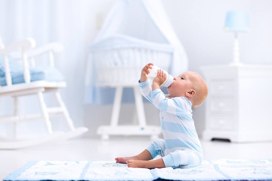 Adorable baby boy playing on a blue floor mat and drinking milk from a bottle in a white sunny nursery with rocking chair and bassinet. Bedroom interior with infant crib. Formula drink for infant.