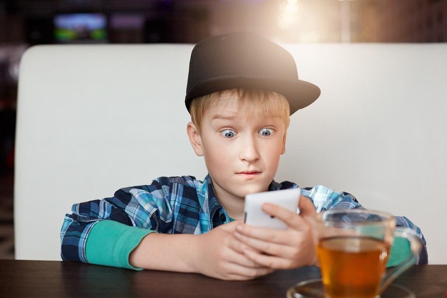 A portrait of little boy wearing modern shirt and cap siting at cafe with a cup of tea holding smartphone looking with great surprise into the screen with wide opened eyes. People and emotions.