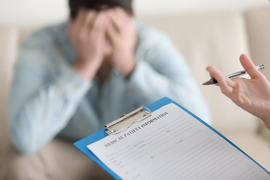 Depressed guy visiting doctor, having complaints about health or mental problems. Focus on doctors hand holding clipboard with patient card, psychologist starts working with troubled man. Close up