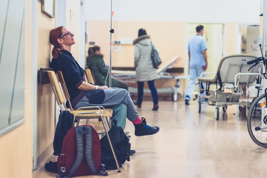 Patient sitting in hospital ward hallway waiting room with iv. Woman with intravenous therapy in her hand is waiting in the clinic corridor with blurred medical personnel in background.